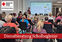 Dienstberatung Schulbegleiter: Eine Gruppe von Personen sitzt in einem Konferenzraum. Eine Frau steht am Tisch und spricht, während ein Projektor eine Präsentation an die Wand wirft..