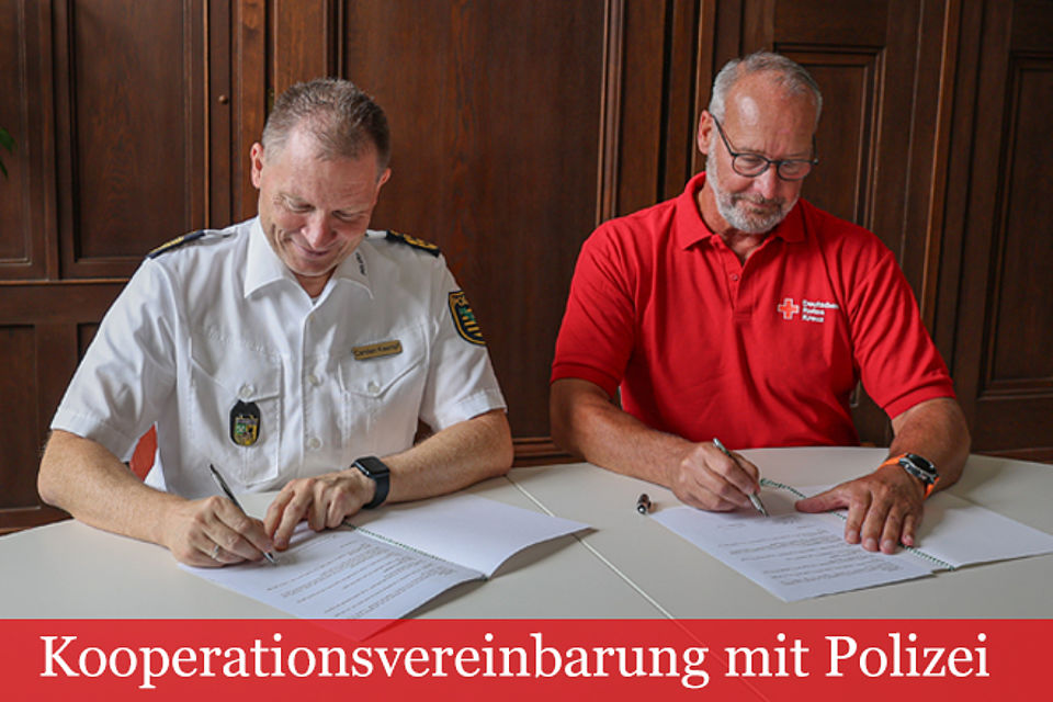 Polizeipräsident Carsten Kaempf im weißen Polizei-Diensthemd und Thomas Illert, Vorstandsvorsitzender des DRK Kreisverbandes Aue-Schwarzenberg e.V. mit rotem DRK T-Shirt, sitzen an einem Tisch und unterzeichnen jeder eine Vereinbarung