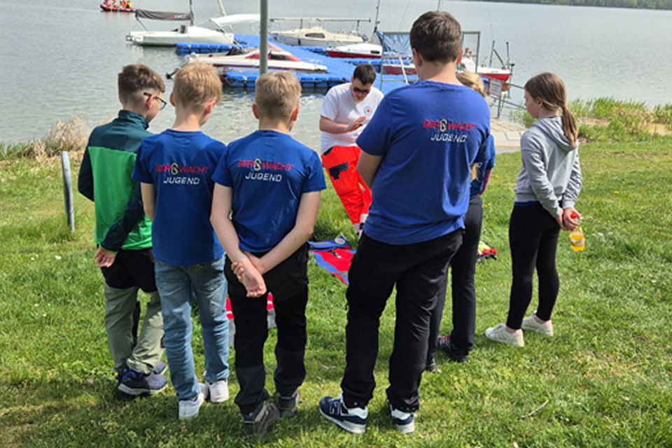 Fünf Kinder und Jugendliche beobachten die Unterweisung eines Kameraden der Wasserwacht.