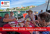 Sommerfest ServiceWohnen: Senioren reichen sich am Kaffeetisch Teller zu, im Hintergrund die Auer Friedenskirche