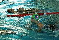Zwei Personen treiben auf dem Rücken im Schwimmbecken, eine weitere Person mit grünem Badekappe und rotem Kreuz schwimmt neben einer Bahnmarkierung