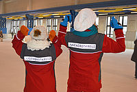 Zwei Jugendliche mit dicker Jacke und Mütze - Aufnahme von hinten. Auf den Jacken ist die Aufschrift JUGENDROTKREUZ und die die Personen zeigen mit den Daumen über die Schulter auf die Aufschrift
