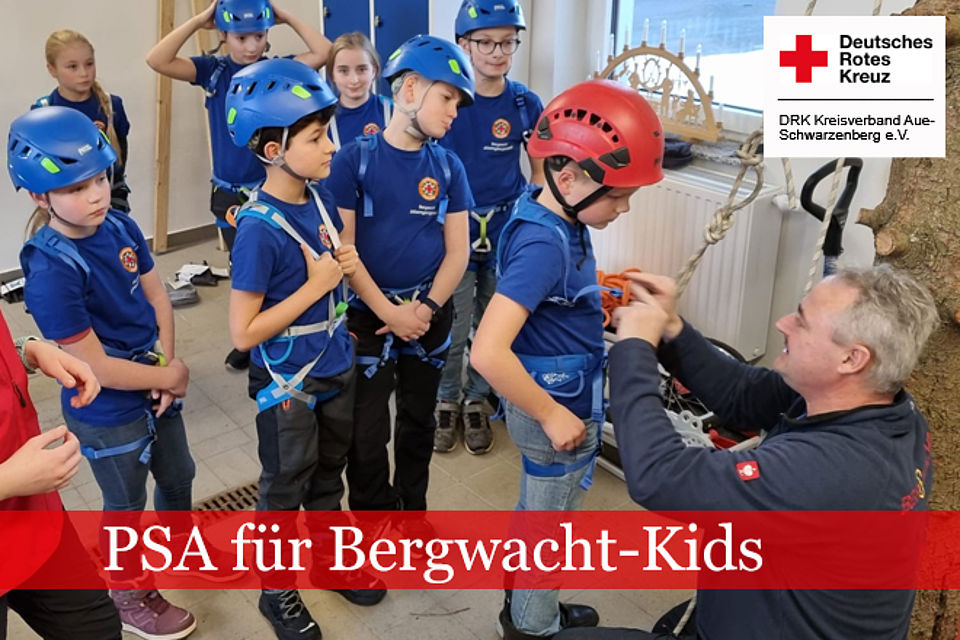 Einsatzkraft der Bergwacht legt einem Kind Schutzausrüstung an. Weitere Kinder stehen dahinter und schauen zu.