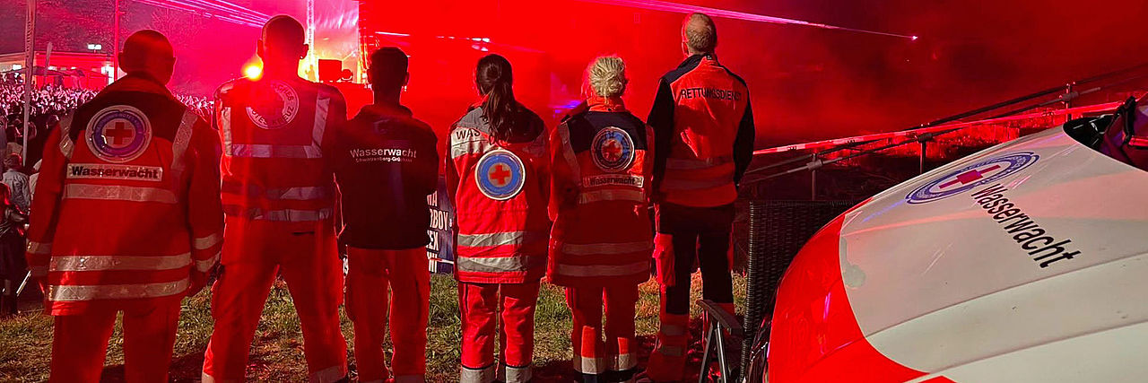 Einsatzkräfte der Wasserwacht in Dienstkleidung stehen neben ihrem Auto und beobachten ein Konzert