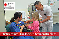 Kindergartenkind legt einer Pflegekraft eine Blutdruckmanschette an und wird von einem Pfleger unterstützt.