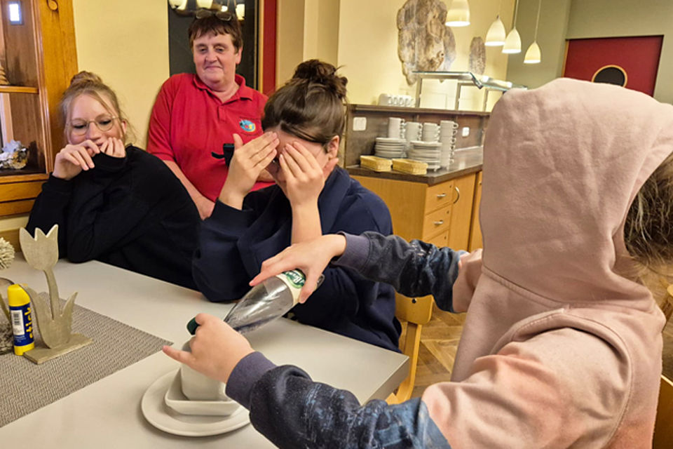 660_2025_JRK_AWE_Herbst_3.jpg Drei Kinder testen, wie man mit verbundenen Augen Wasser in eine Tasse füllt. Einem Kind sind die Ausben verbunden, die anderen beiden schauen konzentriert zu.