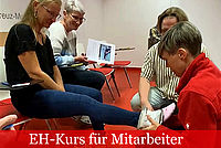 Vier Personen üben Erste Hilfe. Eine Frau in einer roten Jacke hockt vor einer Frau, die auf einem Stuhl sitzt und legt ihr einen Verband am Knöchel an.