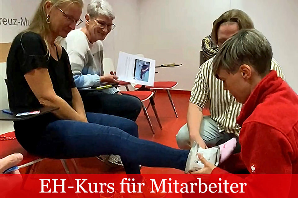 Vier Personen üben Erste Hilfe. Eine Frau in einer roten Jacke hockt vor einer Frau, die auf einem Stuhl sitzt und legt ihr einen Verband am Knöchel an.