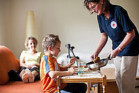 Helferin in DRK Kleidung gibt kleinem Jungen an einem Kindertisch Essen aus einer Bratpfanne auf seinen Teller. Mutter mit verletztem Bein sitzt auf dem Sofa und beobachtet die Aktion