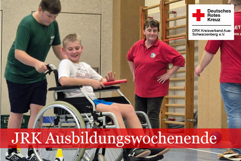 660_2025_JRK_AWE_Herbst_5.jpg JRK Ausbildungswochenende. Ein Junge schiebt einen anderen Jungen im Rollstuhl durch die Turnhalle. Eine Betreuerin in DRK Kleidung schaut zu.
