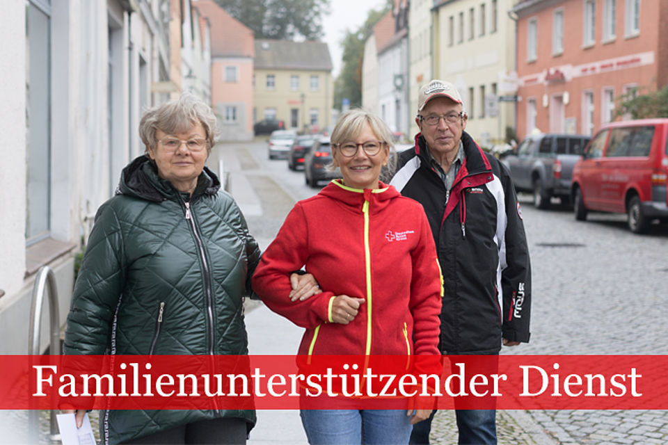 Familienunterstützender Dienst: Mitarbeiterin in DRK Kleidung geht mit einem älteren Ehepaar die Straße entlang. Die hält die Frau eingehenkelt.
