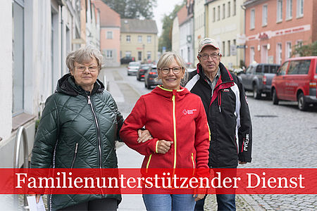 Familienunterstützender Dienst: Mitarbeiterin in DRK Kleidung geht mit einem älteren Ehepaar die Straße entlang. Die hält die Frau eingehenkelt.
