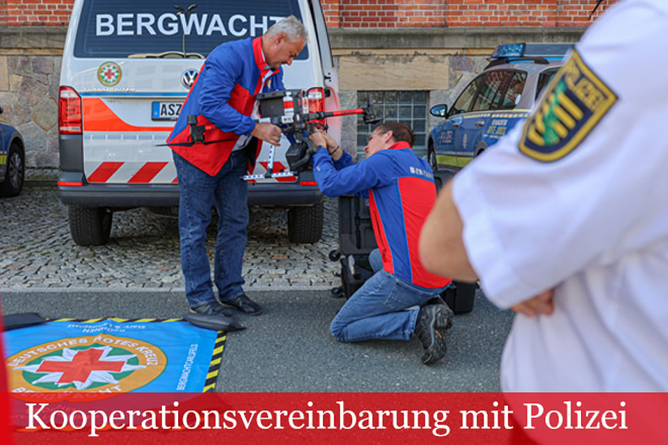Schriftzug Kooperationsvereinbarung mit Polizei: Zwei Angehörige der Bergwacht, einer kniend mit einer Drohne, der andere stehend. Im Hintergrund ein Einsatzfahrzeug der Bergwacht und ein Polizeiauto.