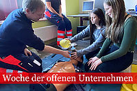 Praxisanleiter in DRK Kleidung erläutert zwei Jugendlichen den Umgang mit einem Beatmungsbeutel. Alle drei knien neber einer Übungspuppe zur HLW.