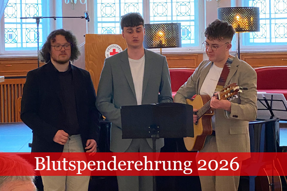 660_2026_Blutspenderehrung_5.jpg Blutspenderehrung 2026: Drei junge Männer stehen hinter einem Notenständer und singen ein Musikstück, die rechte Person hat eine Gitarre.