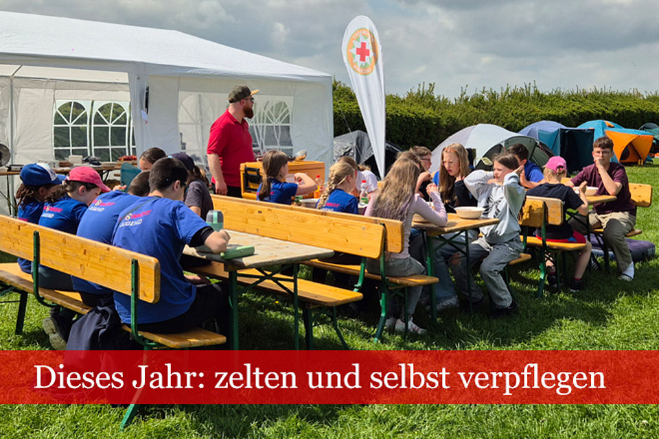 Mehrere Kinder und Jugendliche, einige davon in DRK- oder Bergwacht-Bekleidung sitzen im Freien an Biertischen beim Essen.