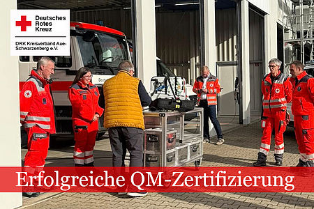 Erfolgreiche QM-Zertifiziereun: Mehrere helfer in DRK-Einsatzkleidung und eine Person mit gelber Weste stehen vor einem DRK-LKW. Auf einem Wagen vor ihnen liegen verschiedene Metallkästen und Arbeitsgeräte.