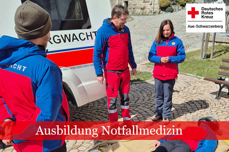 Drei Teilnehmer am Sanitätslehrgang in Bergwachtbekleidung stehen vor einem Bergwachtfahrzeug und schauen auf eine am Boden liegenden Person zur weiteren Versorgung.