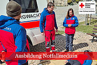 Drei Teilnehmer am Sanitätslehrgang in Bergwachtbekleidung stehen vor einem Bergwachtfahrzeug und schauen auf eine am Boden liegenden Person zur weiteren Versorgung.