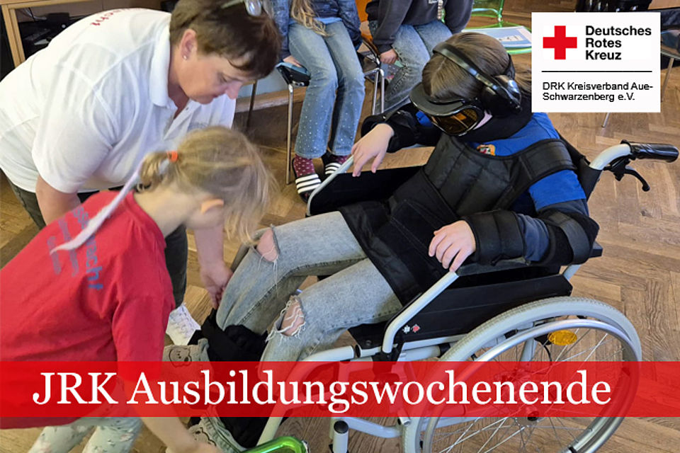 660_2025_JRK_AWE_Herbst_4.jpg JRK Ausbildungswochenende: Ein Mädchen und eine Frau in DRK Kleidung helfen einen Kind beim Anlegen des Alters-Simulationsanzuges. Das Kind sitzt dabei im Rollstuhl.