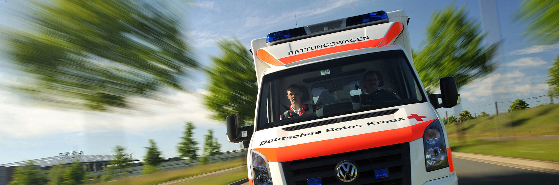 Rettugswagen mit DRK Logo fährt auf der Straße, ein verwaschener Hintergrund suggeriert eine schnelle Fahrt
