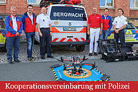 Flugdrohne der Bergwacht auf einem blauen Landekissen mit Bergwacht-Logo, dahinter stehen drei Angehörige der Bergwacht, zwei Vertreter der Polizei und ein Mitglied des DRK, im Hintergrund je ein Fahrzeug von Bergwacht und Polizei