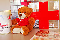 Brauner Teddybär mit rotem T-Shirt sitzt neben einer Spendenbüchse und einem großen roten Kreuz