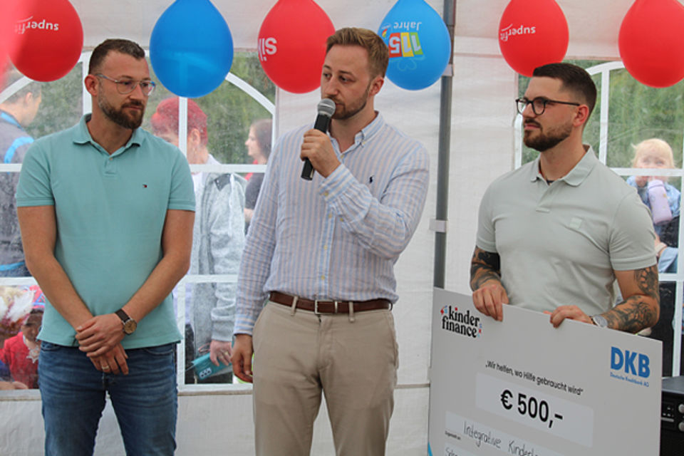 Drei Männe rin einem Zelt mit Luftballons, einer hält einen großen Spendenscheck in der Hand