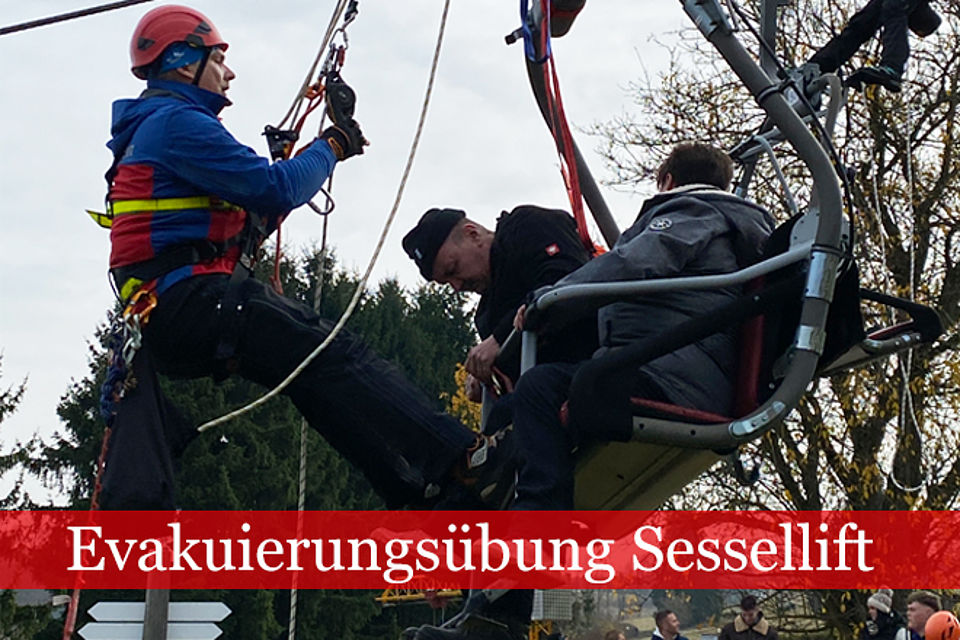 Ein Retter in Bergwachtkleidung seilt sich zu zwei Personen ab, die im Sessellift sitzen